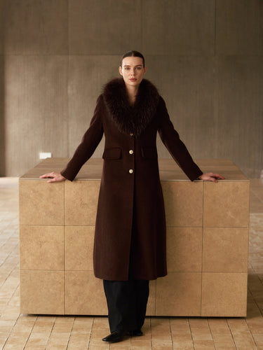 Full-length front view of the Chocolate Brown long cashmere coat with fur collar, gold-tone buttons and a shaped waist, worn over black trousers.