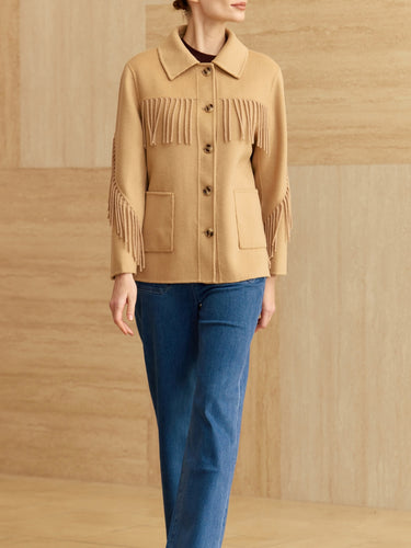 Model wearing a camel jacket with a pointed collar, button front, patch pockets, and fringe across the chest and sleeves, paired with blue jeans.