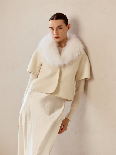 Model wearing the Soft Ivory short-sleeve cashmere jacket with a white fur collar over an ivory knit and satin midi skirt.