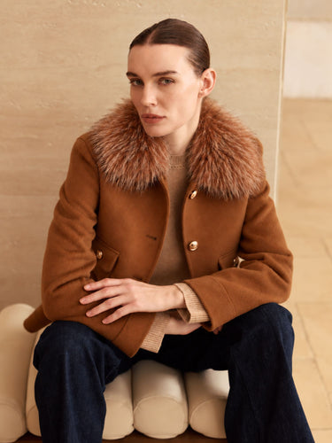 Seated view of the Warm Camel cashmere bomber jacket, showing the full fur collar, gold-tone buttons and buttoned flap pockets.